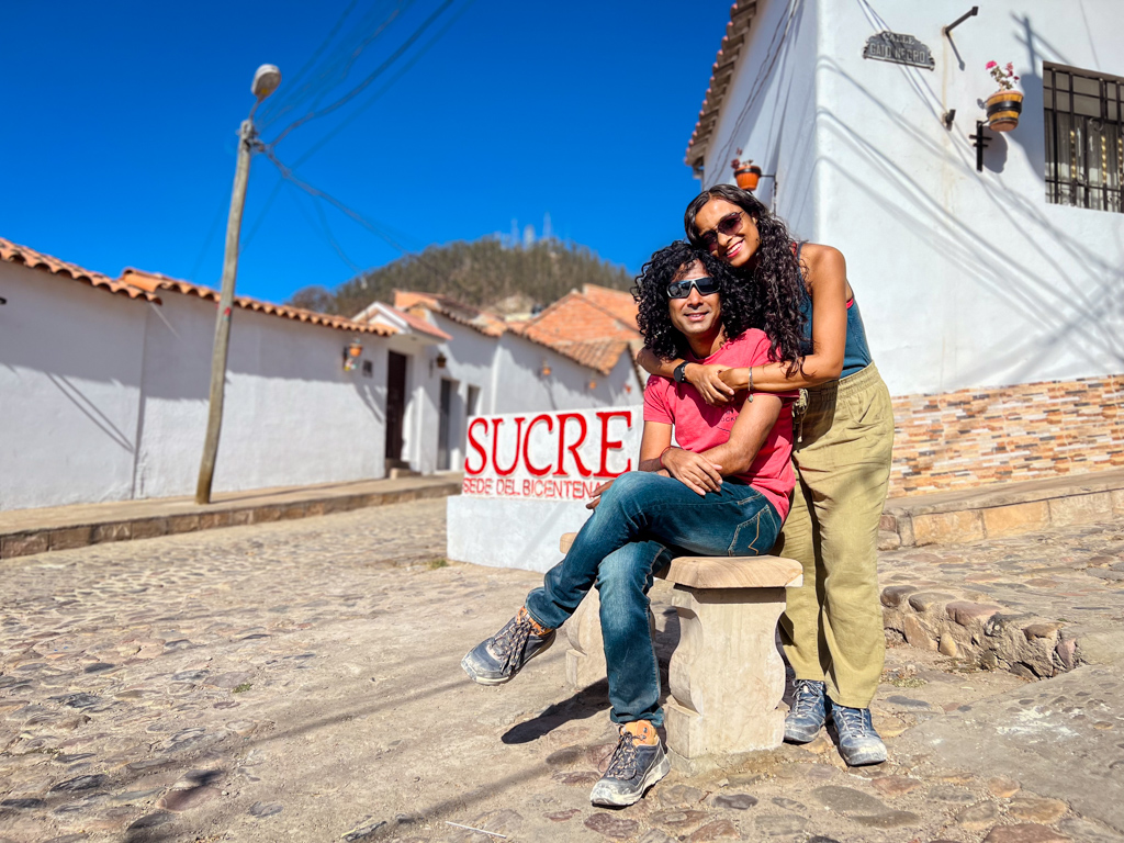 A man sitting and a woman standing on a cobbled street with white houses with terracotta roofs behind in Sucre, the capital of Bolivia 