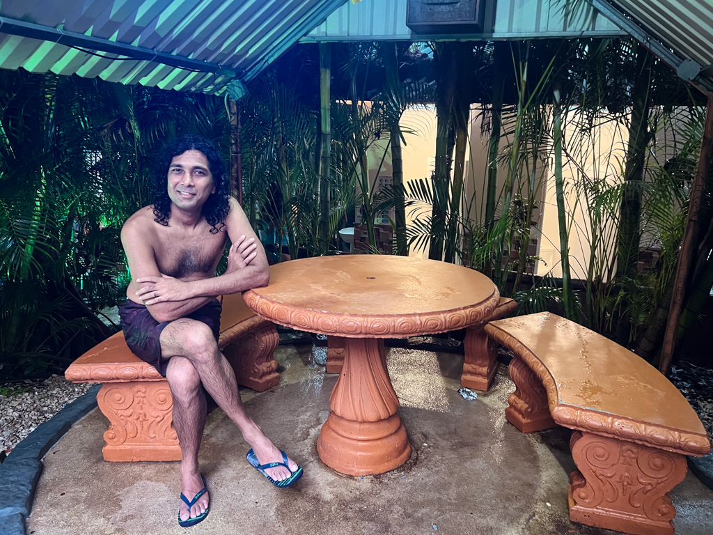 Man seated on a bench next to a table, under the shade of a hut like structure.