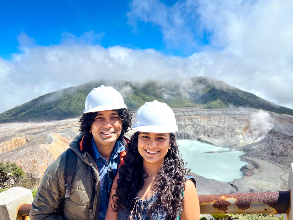 A woman and a man, wearing white helmets, with the crater lake of Poas Volcano behind, in Costa Rica.