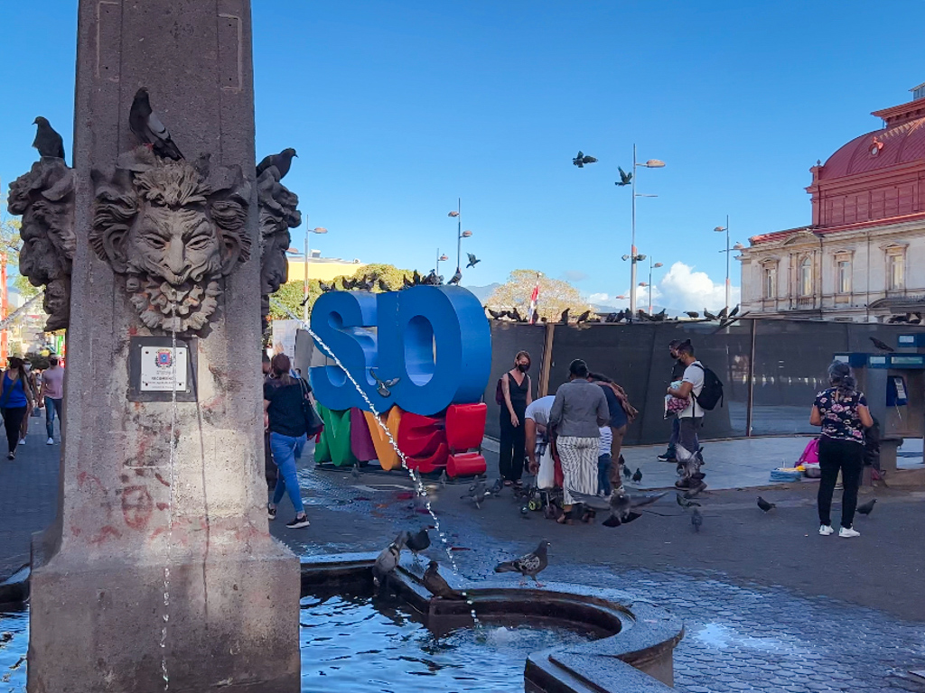 A fountain where water is flowing from statue faces. There is SJO VIVE sign behind and people walking by in the central part of San Jose, Costa Rica.