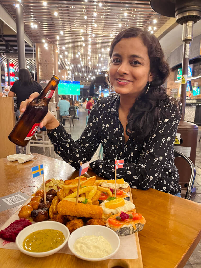 A woman with a beer bottle in hand in a food market with a dish of fried food and sushi on the table in a mall in San Jose, Costa Rica.