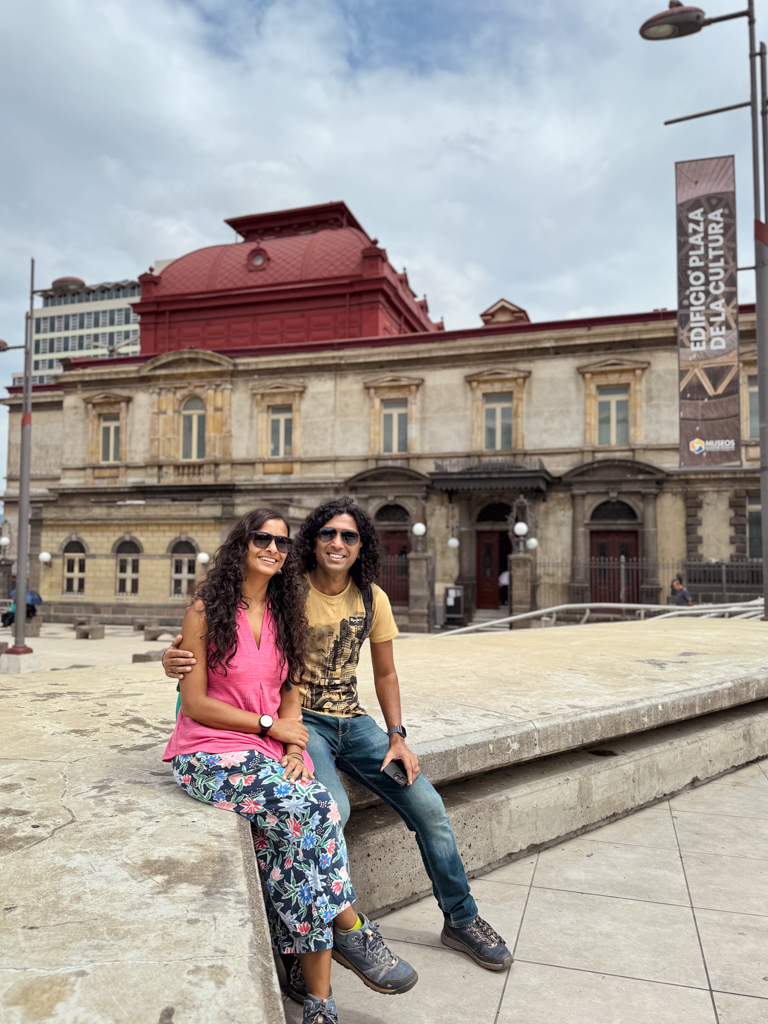 A man and a woman sitting in a plaza at the center of San Jose, Costa Rica. Behind them, there is the National Theater building.