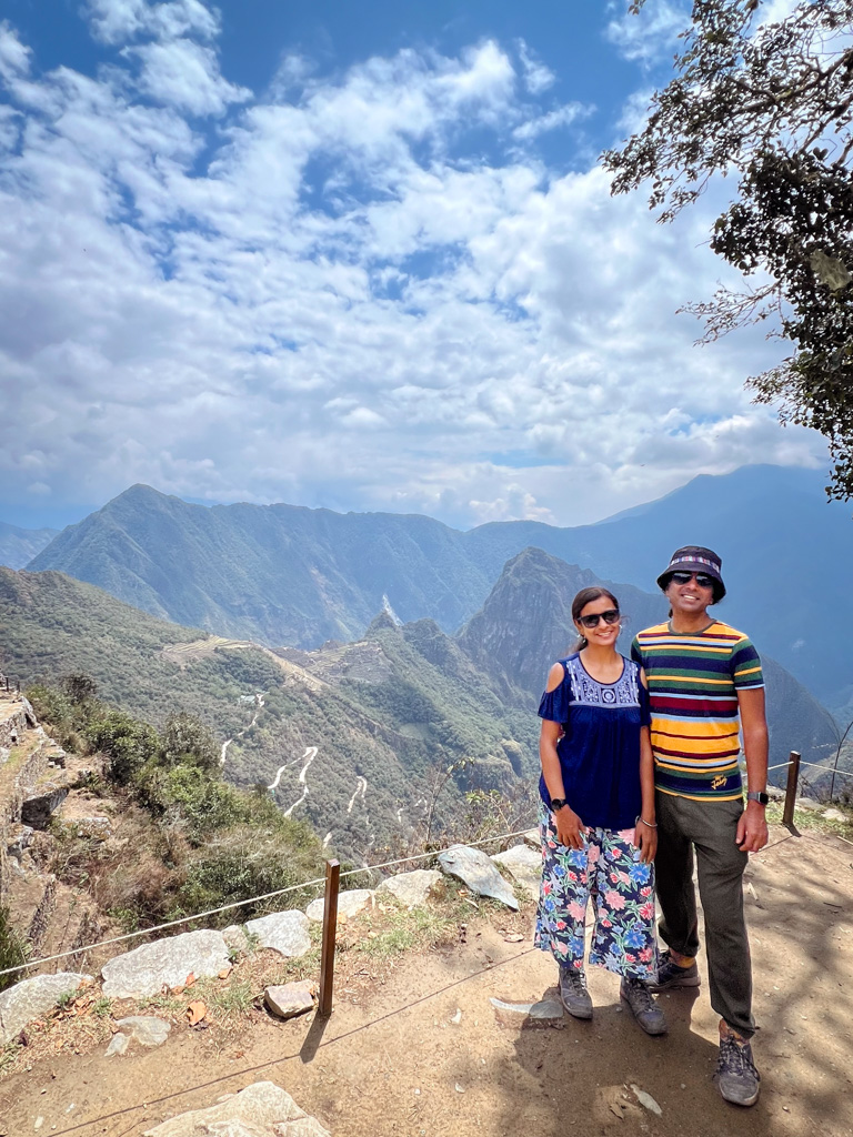 Couple, posing for a photo, at the edge of Inti Punku Sun Gate with Machu Picchu view in the background.