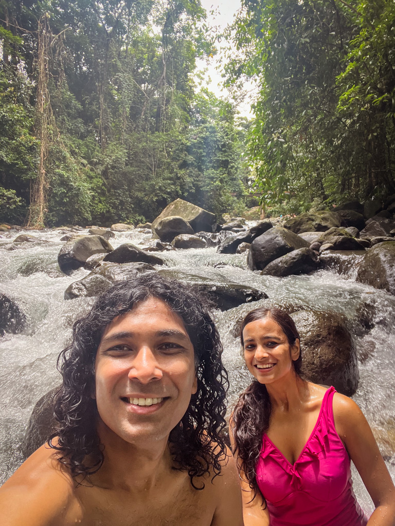 A man and a woman in the Fortuna River in La Fortuna, with a forest behind.