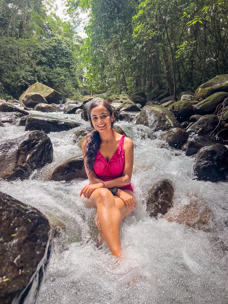 A woman sitting on a large rock in Fortuna river near La Fortuna in Costa Rica. The river water flowing around her, and it is in a forest setting with lots of trees and more rocks in the background. Free Things to Do in La Fortuna.